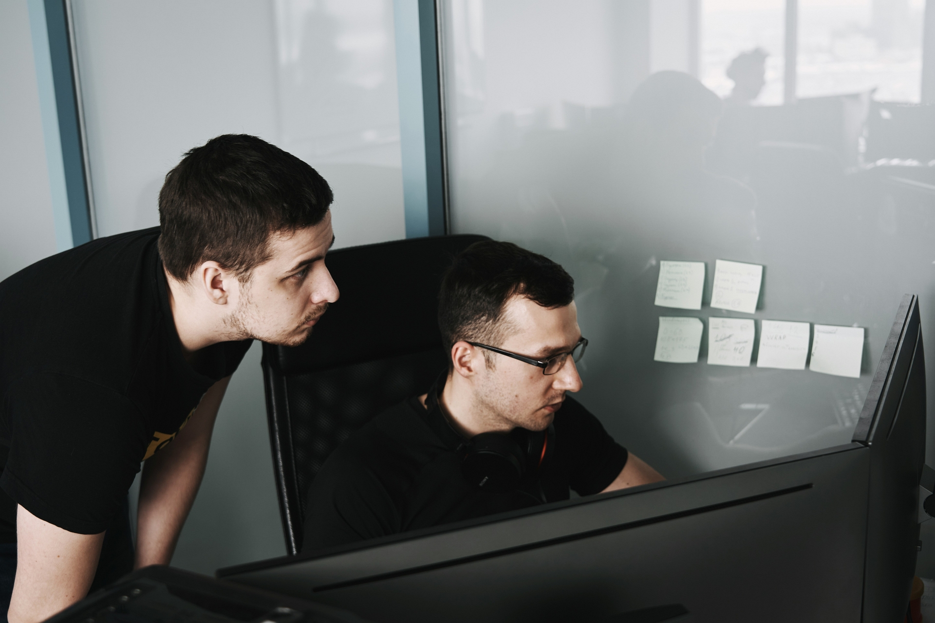 Two guys looking at computer monitors; one hovering over chair while the other sits