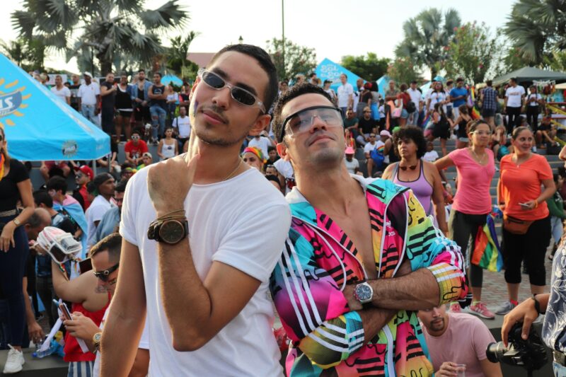 Crowded outdoor event, two happy guys with sunglasses looking at camera, one making hand gestures and the other with arms folded