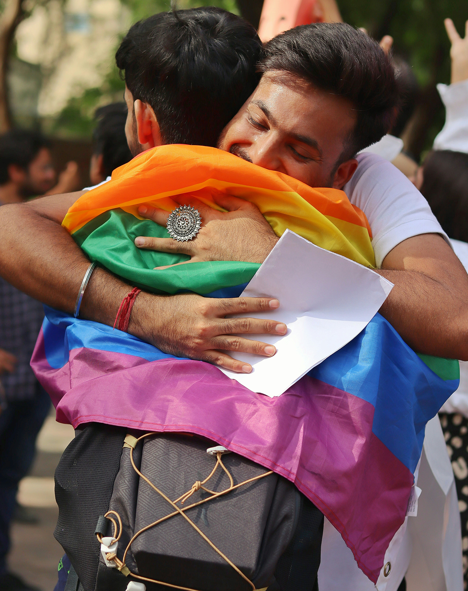 Two guys hugging, one holding apiece of paper and the other wearing a rainbow flag